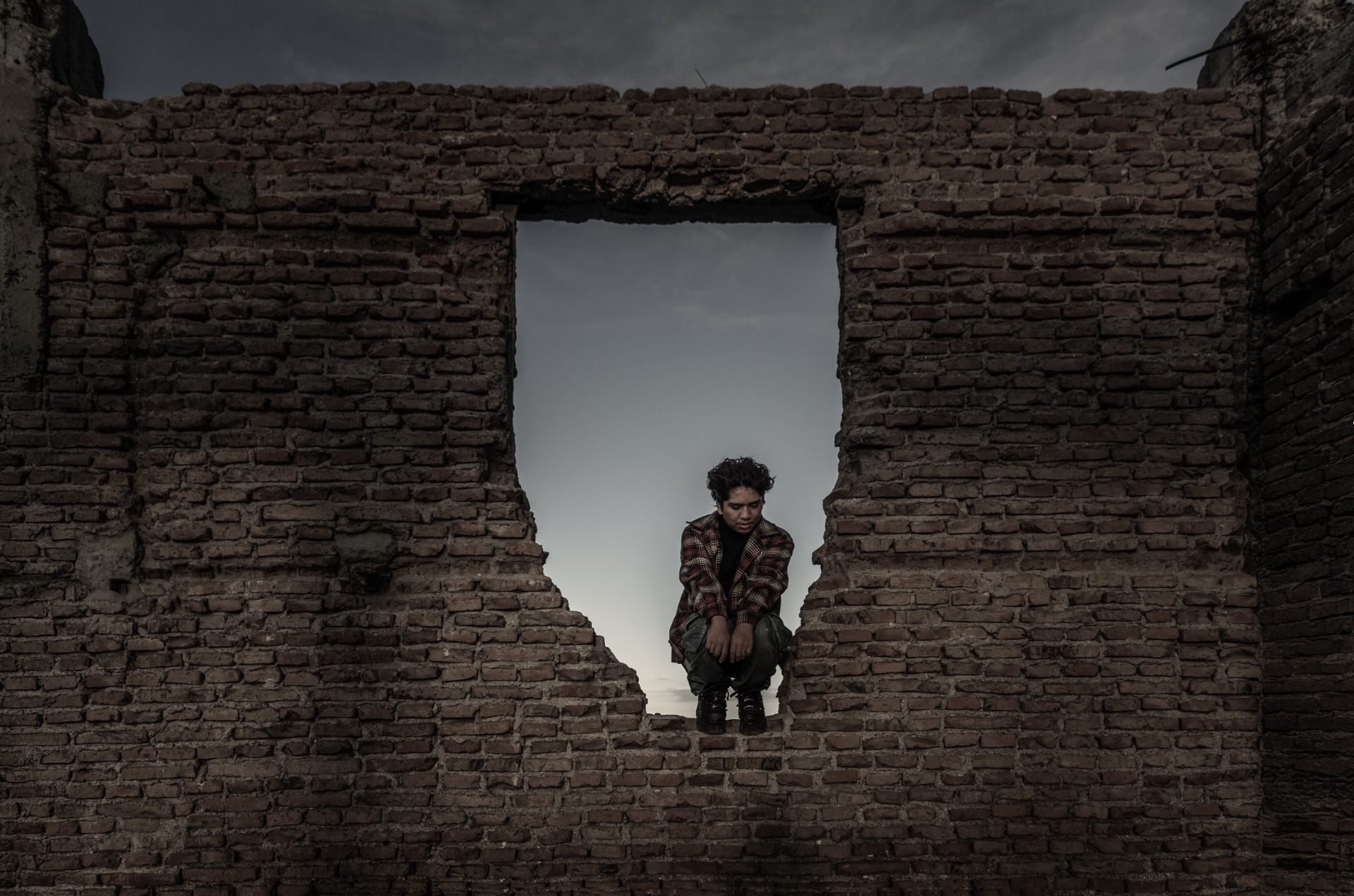 Joven cabizbajo sentado entre ruinas de ladrillo, mirando al suelo bajo cielo nublado.